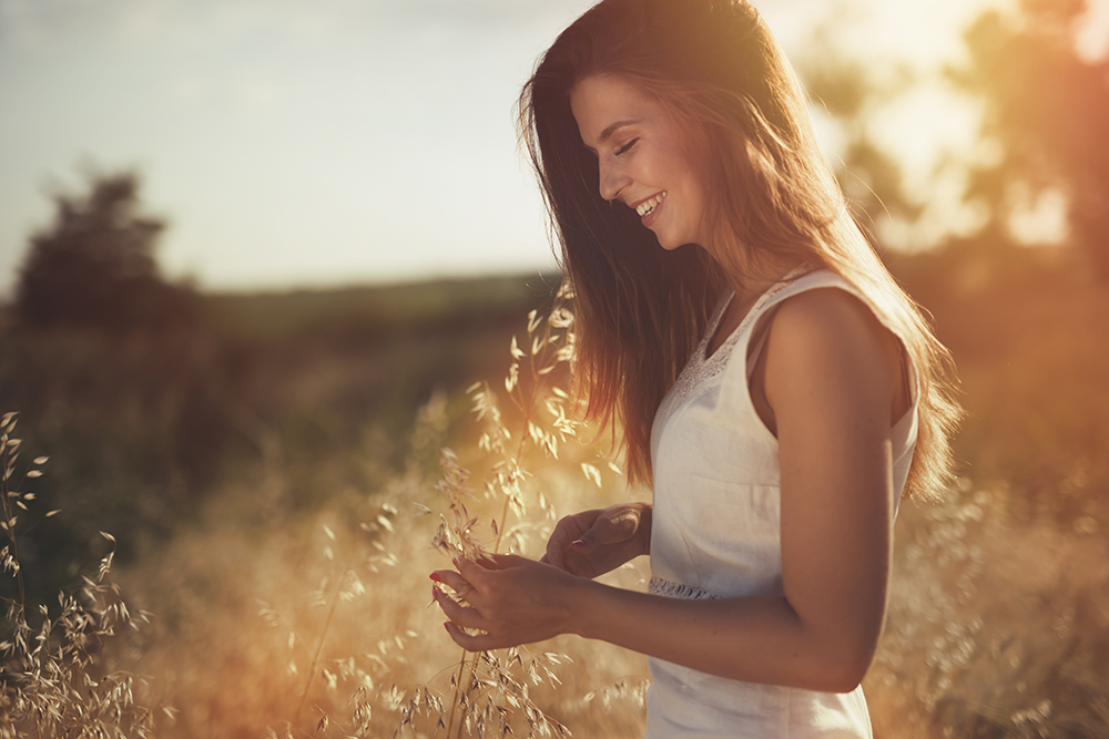 A young woman in a wheat field at golden hour, smiling down at and holding a piece of wheat grass