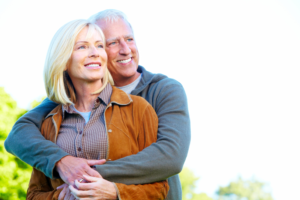 older couple outside smiling as husband hugs wife from behind her